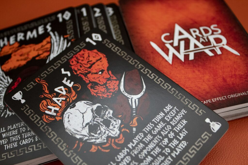 A deck of Cards At War cut in half, with both halves side-by-side. The Hades card is placed across the two halves.